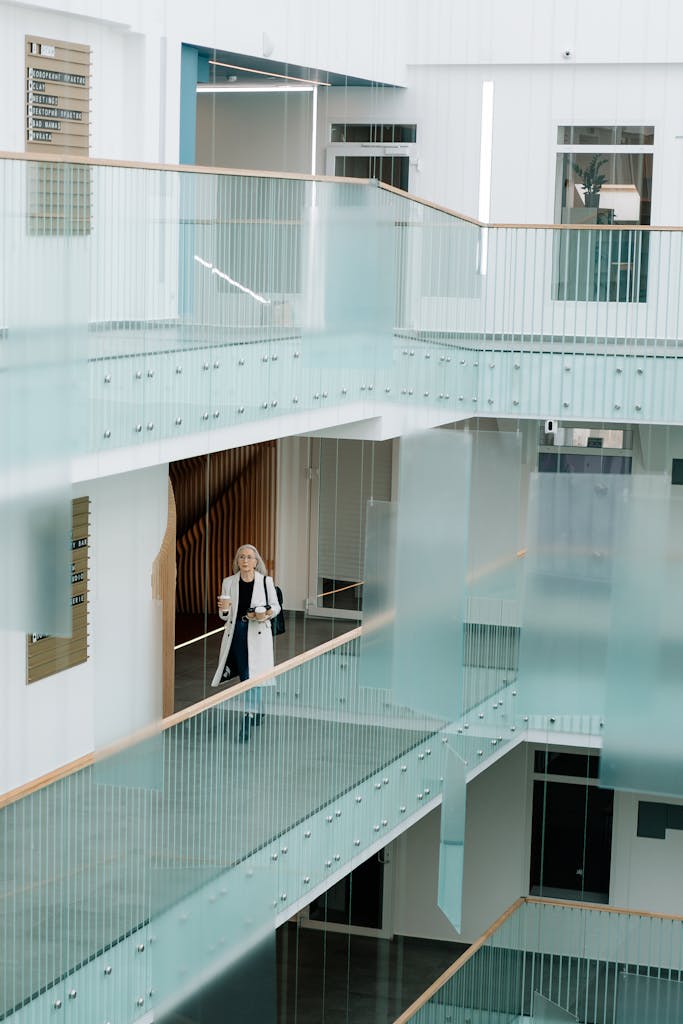 Woman walking through a bright, modern office corridor with glass railings, holding coffee and documents.